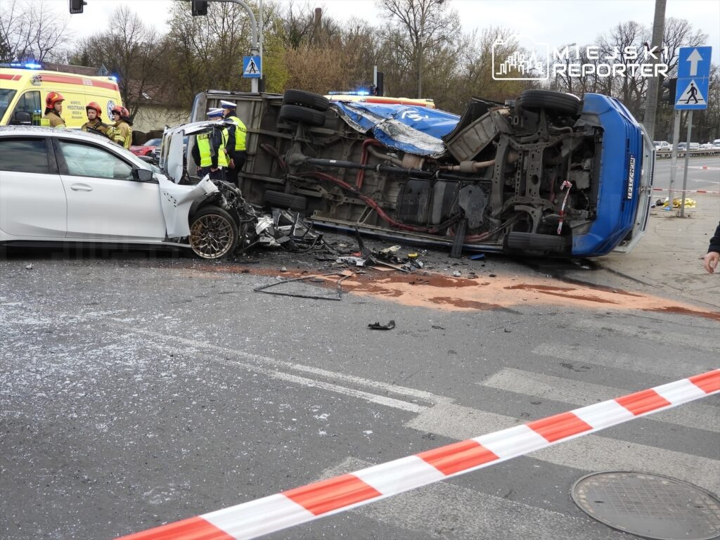 Zniszczony samochód osobowy z uszkodzoną przednią częścią stoi obok przewróconego pojazdu dostawczego. Na miejscu wypadku pracują służby ratunkowe.