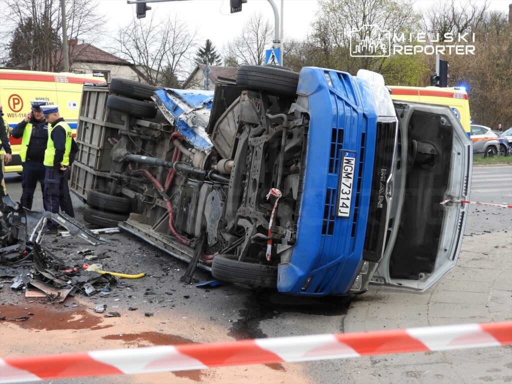Nieoznakowany niebieski pojazd dostawczy leży na boku na jezdni, otoczony przez policjantów i samochody ratunkowe.