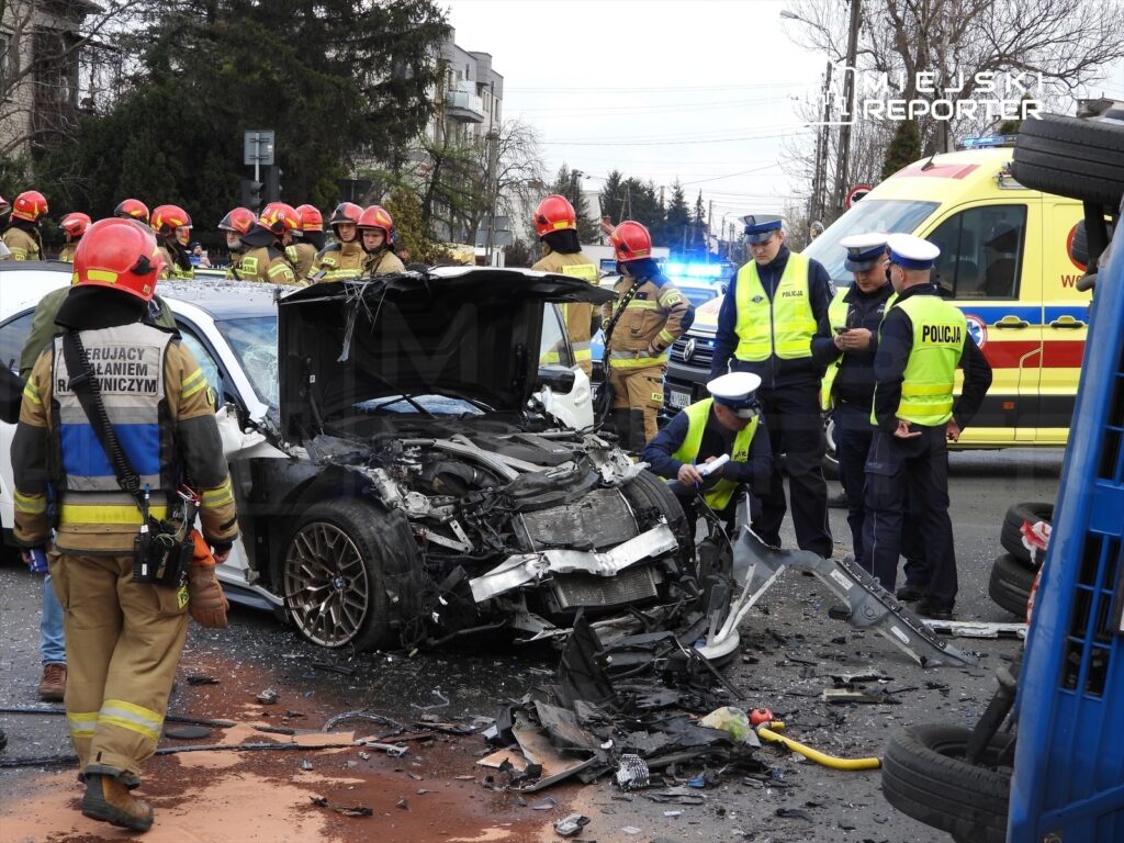 Grupa strażaków i policjantów pracuje przy zniszczonym pojeździe po wypadku drogowym, obok wzywana karetka.