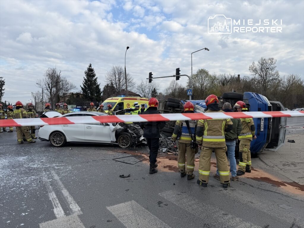 Na skrzyżowaniu leży przewrócony niebieski pojazd, a obok niego biała limuzyna z uszkodzoną przednią częścią. Grupa strażaków w hełmach i mundurach zabezpiecza miejsce zdarzenia, a w tle widoczne są radiowozy i karetka.