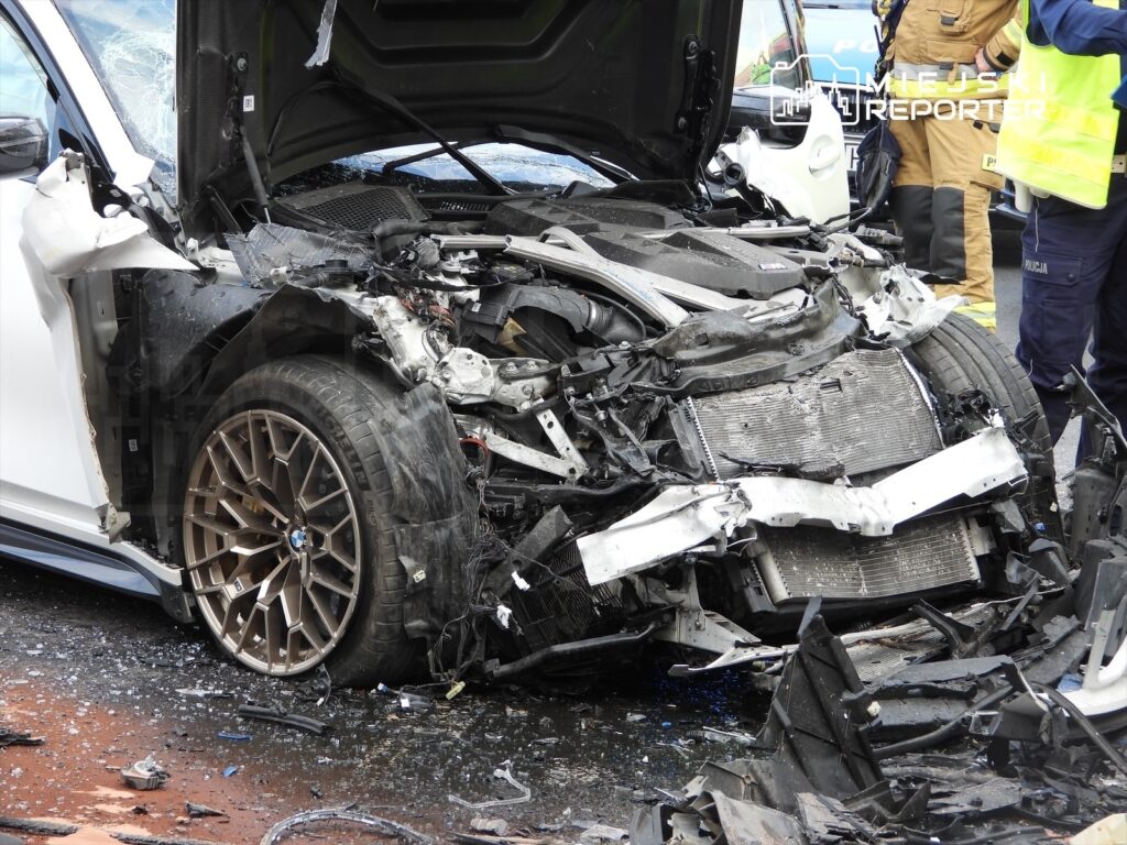 Zniszczony samochód sportowy z odczepioną maską i uszkodzoną przednią częścią, otoczony przez służby ratunkowe.