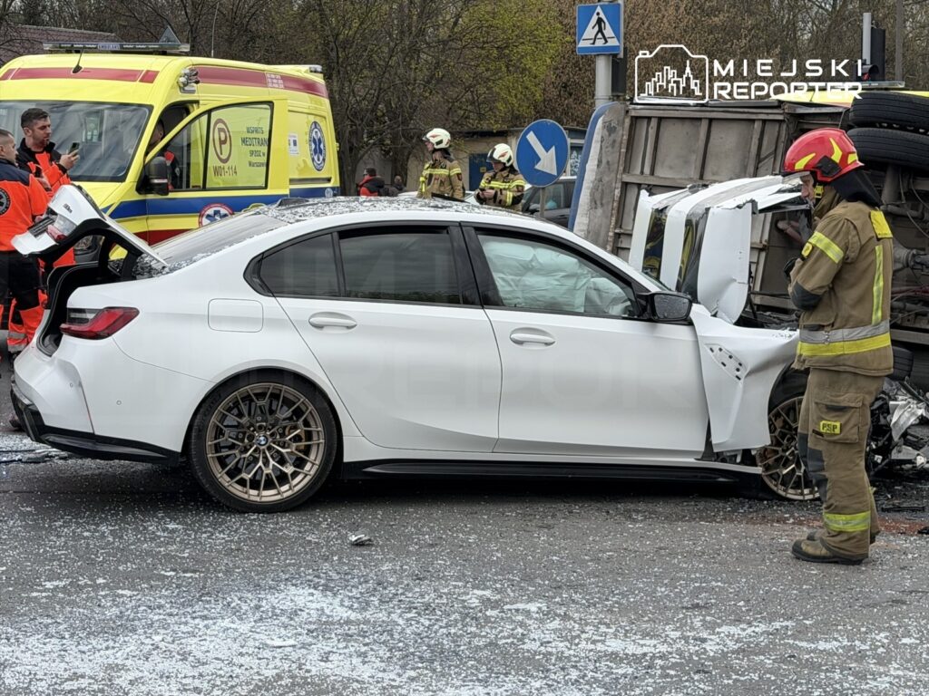 Biały samochód osobowy z uszkodzoną karoserią stoi obok przewróconego pojazdu ciężarowego, w otoczeniu strażaków i ratowników medycznych.