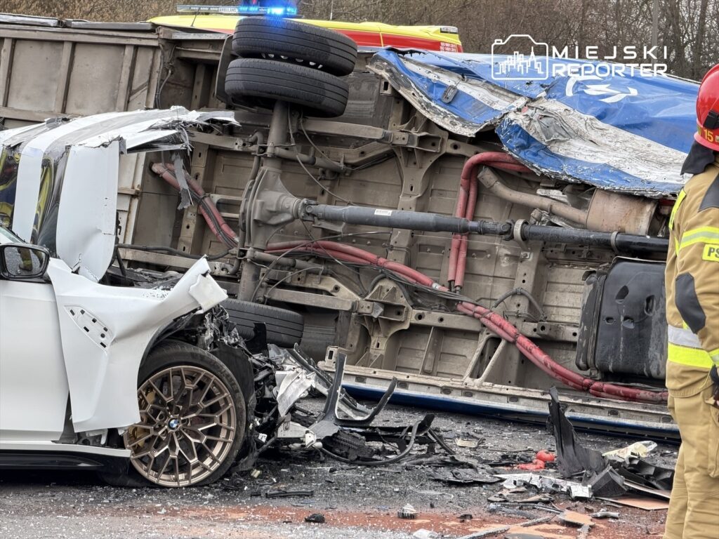W wyniku wypadku na drodze leży przewrócony ciężarowy pojazd, obok uszkodzony samochód osobowy. W tle widać strażaka w uniformie.