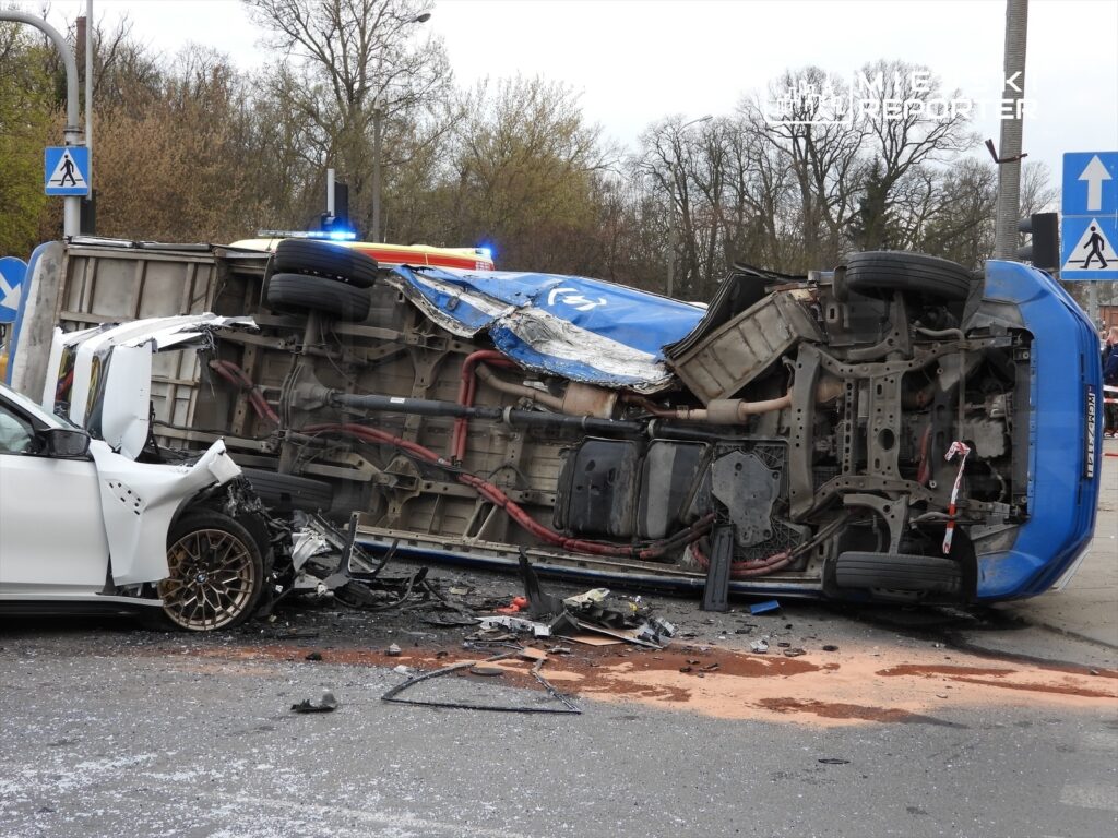 W wyniku wypadku drogowego, niebieski pojazd leży na boku, a obok niego uszkodzony biały samochód. Na miejscu pracują służby ratunkowe.