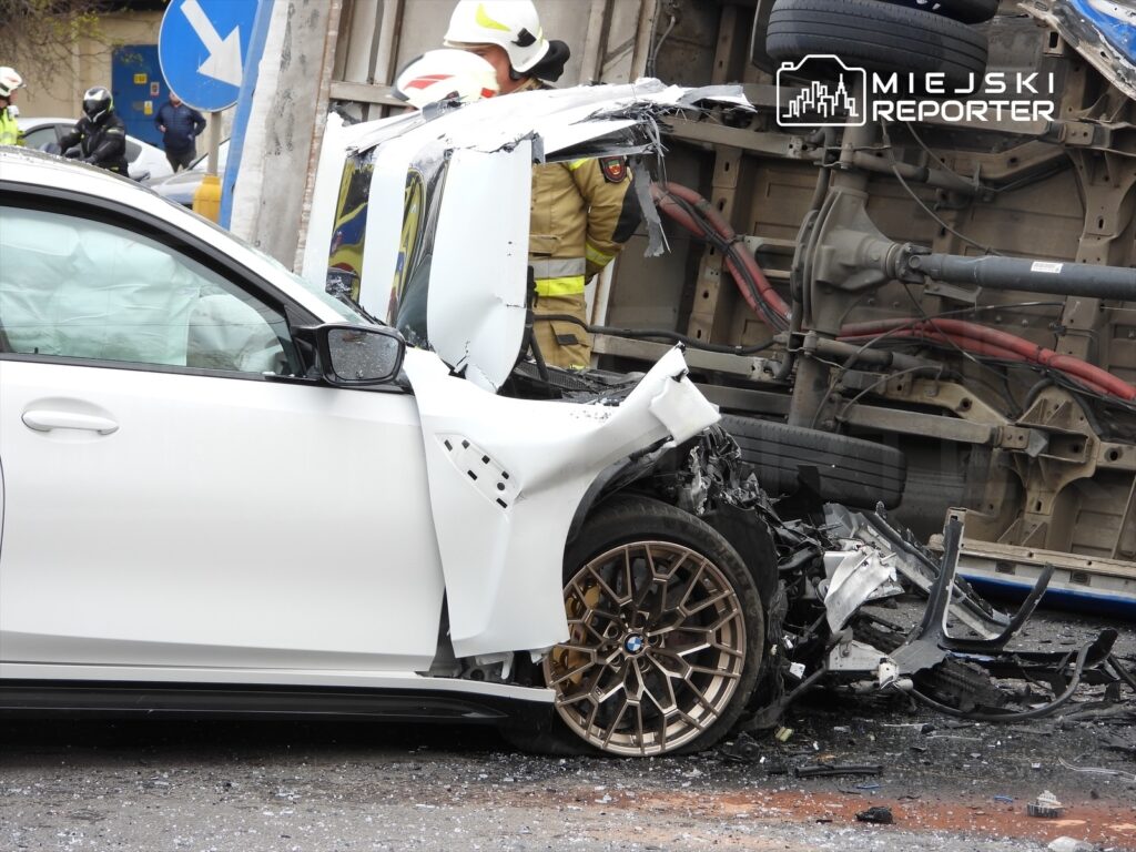 W wyniku wypadku samochodowego zniszczona biała limuzyna stoi obok przewróconego pojazdu dostawczego, w tle strażacy udzielają pomocy.