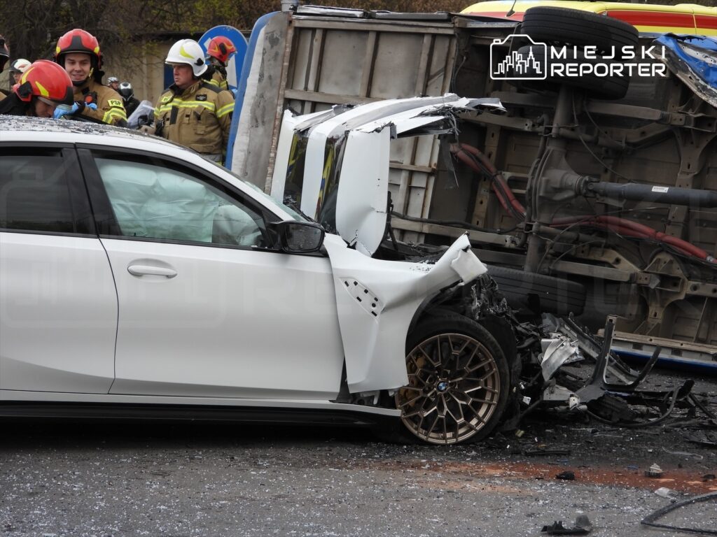 Biały samochód osobowy z uszkodzoną boczną częścią stoi obok przewróconej ciężarówki, a ratownicy udzielają pomocy.