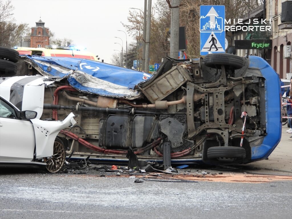 Zniszczony niebieski pojazd leży na boku na jezdni, obok białego samochodu, w tle widać sygnalizację świetlną i budynki.