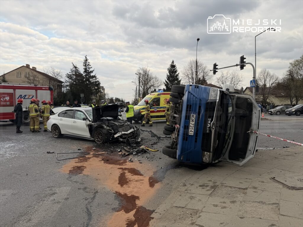 W miejscu wypadku drogowego, uszkodzone białe auto osobowe z otwartą maską oraz przewrócony niebieski pojazd dostawczy. W tle strażacy i policjanci prowadzą działania ratunkowe.