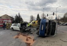W miejscu wypadku drogowego, uszkodzone białe auto osobowe z otwartą maską oraz przewrócony niebieski pojazd dostawczy. W tle strażacy i policjanci prowadzą działania ratunkowe.