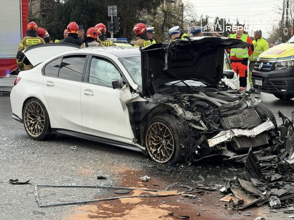 Uszkodzony biały samochód osobowy z otwartą maską stoi na jezdni, a obok niego pracują strażacy w mundurach.