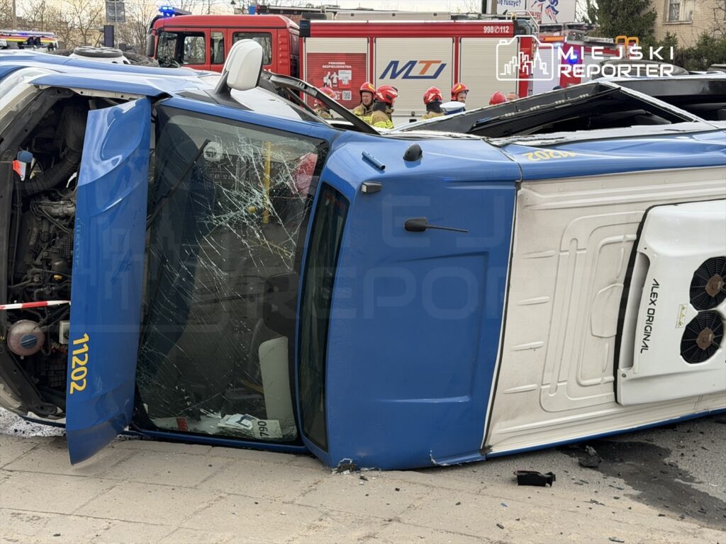 Niebieski samochód dostawczy leży na boku na jezdni, z wgniecioną szybą i otwartymi drzwiami, obok strażacy w mundurach.