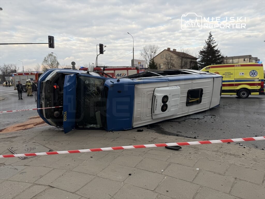 Niebieski pojazd leżący na boku na jezdni, otoczony przez służby ratunkowe i taśmy zabezpieczające.