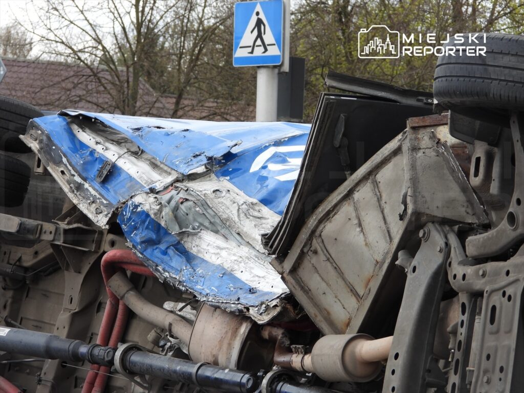 Zniszczony niebieski samochód leży na dachu, odsłaniając uszkodzone podwozie i elementy mechaniczne. W tle widoczny znak drogowy.