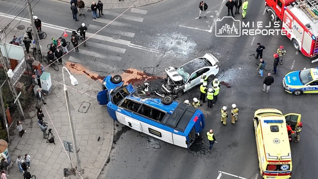 Zniszczony autobus leży na boku obok rozbitego samochodu osobowego, a na miejscu zdarzenia pracują służby ratunkowe.