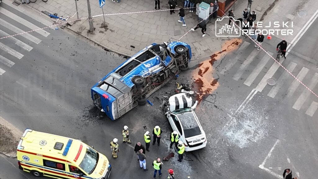 W wyniku wypadku przewrócony niebieski autobus leży na boku, a obok niego uszkodzone białe auto. Ratownicy i policjanci zabezpieczają miejsce zdarzenia, na jezdni widoczne plamy płynów.