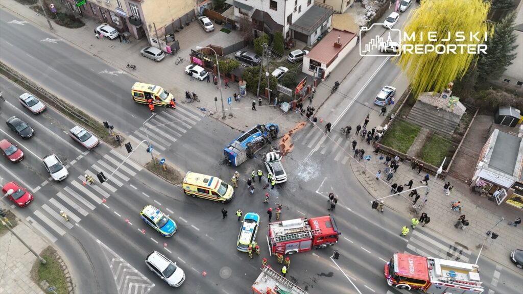 Wielka awaria drogowa z przewróconym niebieskim pojazdem na skrzyżowaniu, otoczona przez ratowników i policję.