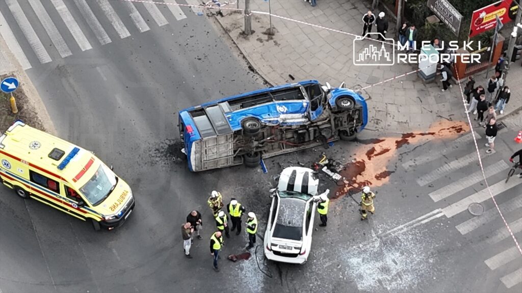 Wielki niebieski autobus przewrócony na bok na skrzyżowaniu, obok grupy ratowników medycznych i strażaków.