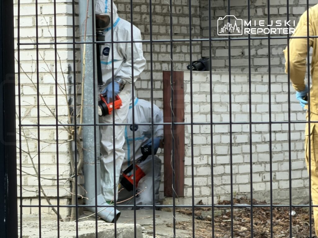 Dwaj mężczyźni w kombinezonach ochronnych pracują przy ścianie budynku, używając narzędzi do usuwania materiałów.