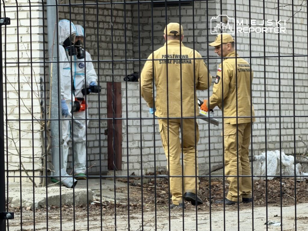 Mężczyzna w specjalnym kombinezonie ochronnym stoi obok dwóch strażaków w mundurach, rozmawiających w pobliżu budynku.