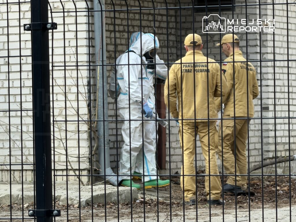 Osoba w kombinezonie ochronnym bada ścianę budynku, a obok stoją strażacy w żółtych uniformach.