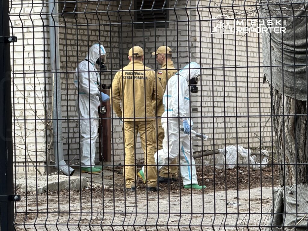 Czterech mężczyzn w kombinezonach ochronnych i mundurach strażackich przebywa w pobliżu budynku, badając okolice.