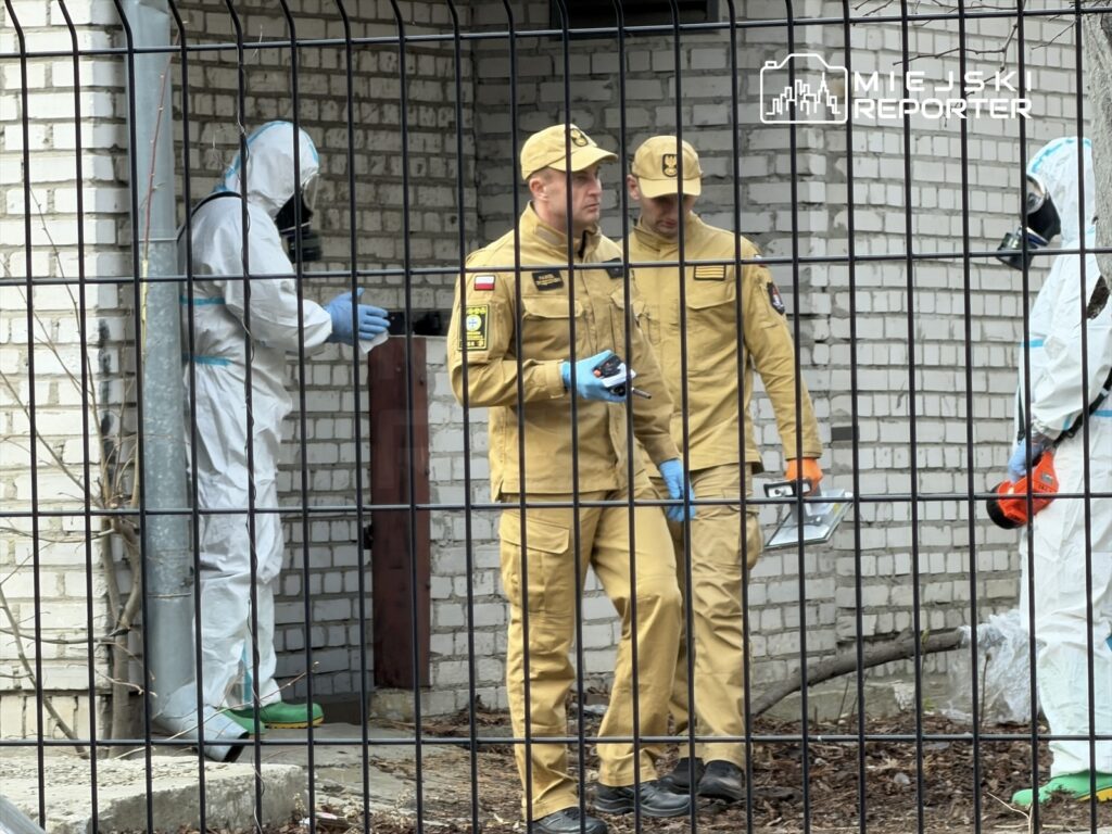 Dwóch mężczyzn w beżowych mundurach omawia sytuację przed budynkiem, podczas gdy osoba w kombinezonie ochronnym pracuje w tle.