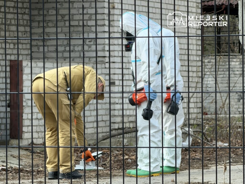 Mężczyzna w kombinezonie ochronnym i drugi w mundurze badają teren przy ogrodzeniu, obok nieużytków.