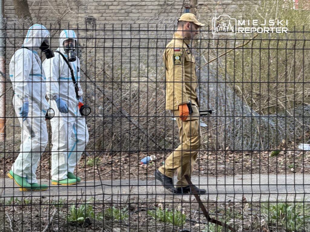 Dwaj mężczyźni w kombinezonach ochronnych przechodzą obok ogrodzenia, a obok nich idzie mężczyzna w mundurze.