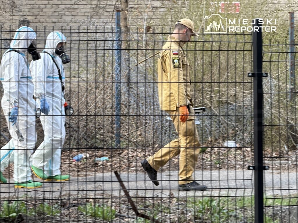 Grupa dwóch osób w białych kombinezonach ochronnych idzie wzdłuż ogrodzenia, podczas gdy mężczyzna w beżowym mundurze trzyma narzędzie.