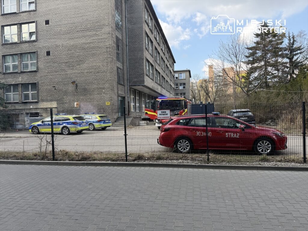 Trzy radiowozy policyjne i wóz strażacki zaparkowane przed budynkiem, w tle widoczne drzewa i chmury.