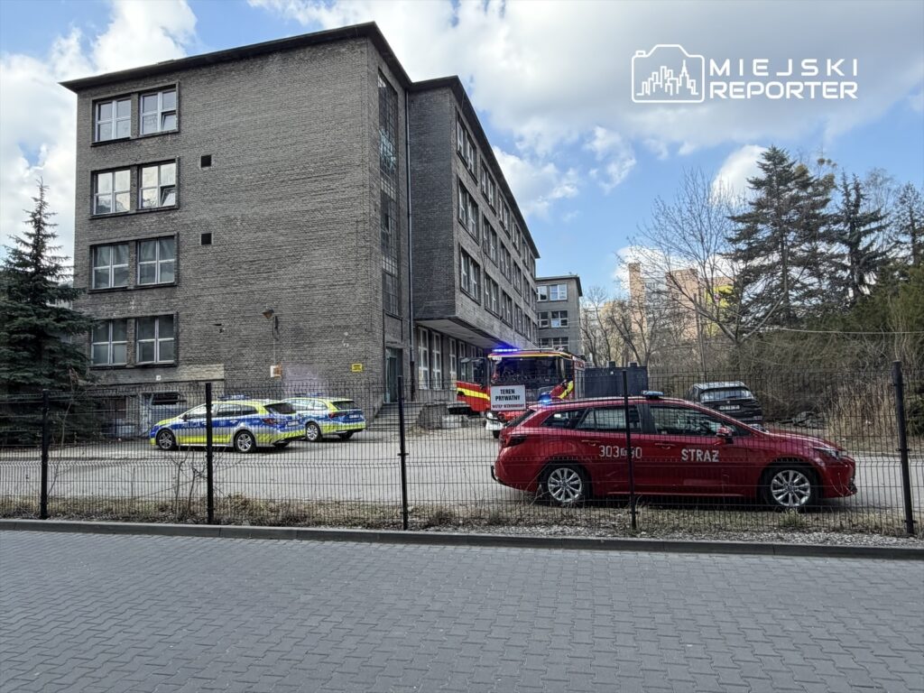 Na podjeździe przed budynkiem stoi czerwona strażacka karetka oraz dwa radiowozy policyjne. W tle widoczne są drzewa i chmury.