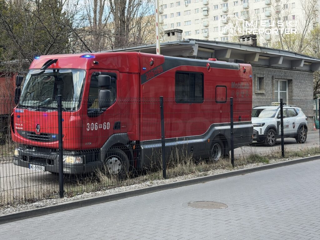 Czerwony pojazd ratunkowy zaparkowany obok budynku mieszkalnego, z włączonymi światłami sygnalizacyjnymi.