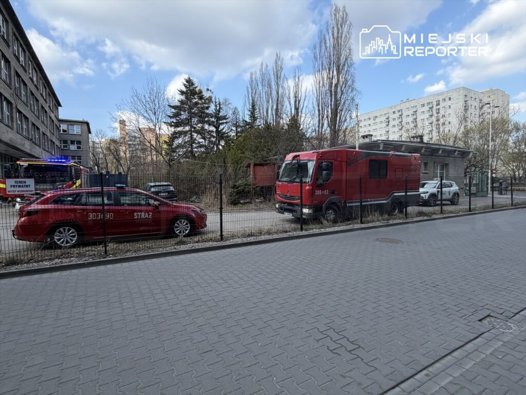 Czerwony samochód strażacki z włączonymi sygnałami świetlnymi stoi obok dużego pojazdu ratunkowego na terenie otoczonym siatką.