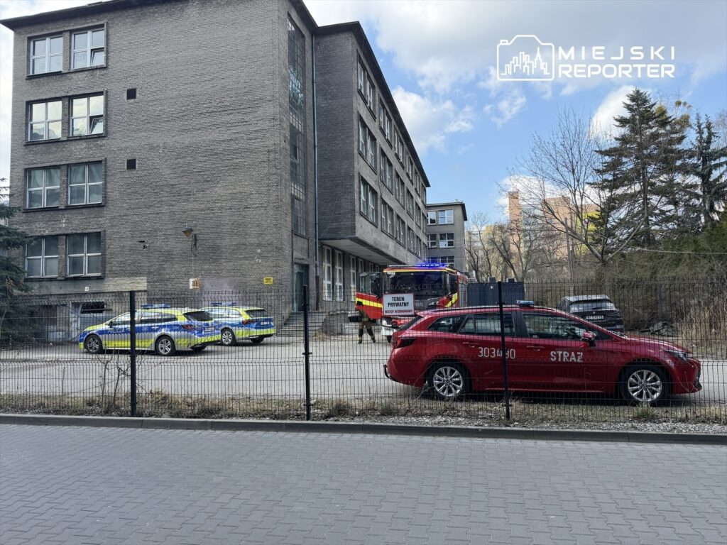 Na placu przed budynkiem stoją policyjne i strażackie pojazdy, w tym czerwony samochód straży pożarnej z włączonymi sygnałami.