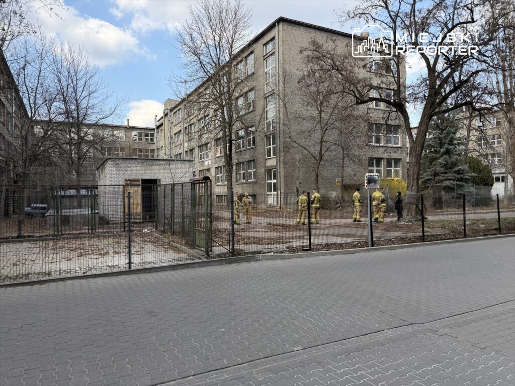 Grupa strażaków w żółtych mundurach przeprowadza działania na terenie otoczonym ogrodzeniem przy szarej, wielopiętrowej kamienicy.