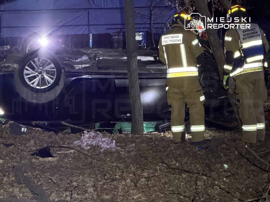 Strażacy w mundurach zabezpieczają przewrócone auto w lesie, obok leżą fragmenty materiałów.