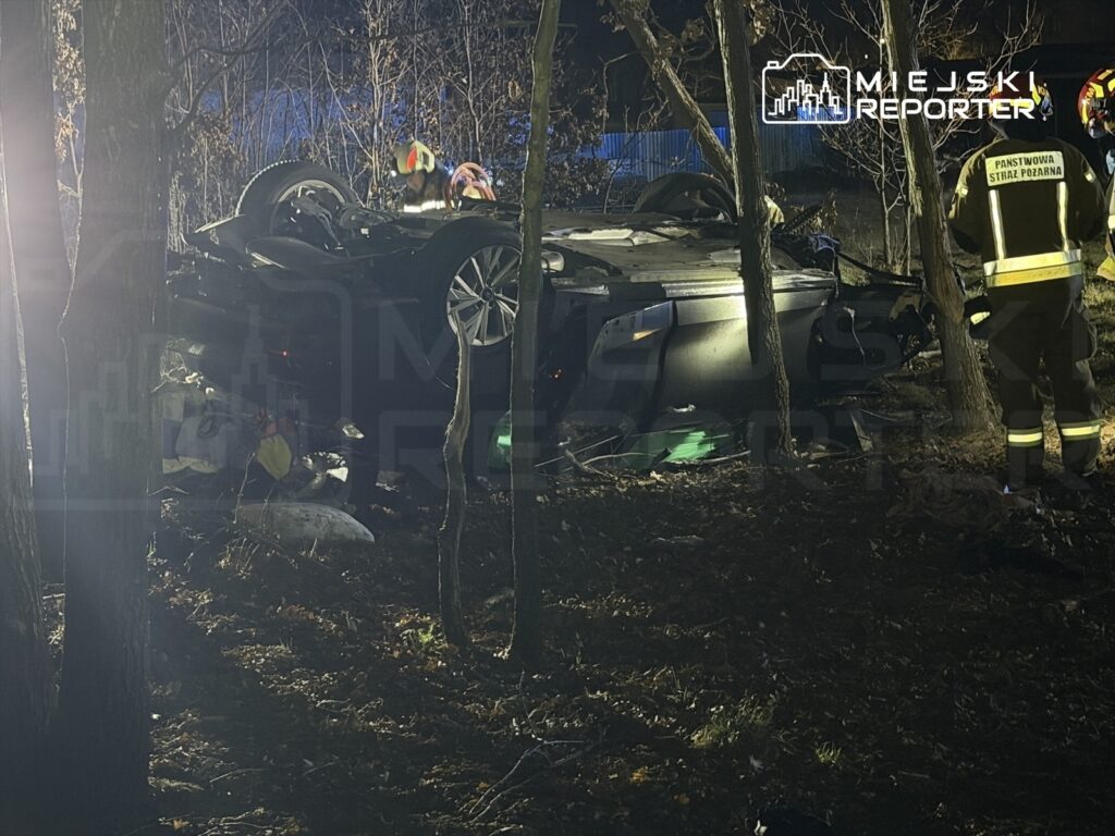 Samochód osobowy przewrócony na dach w lesie, obok strażacy w sprzęcie ochronnym prowadzący działania ratunkowe.
