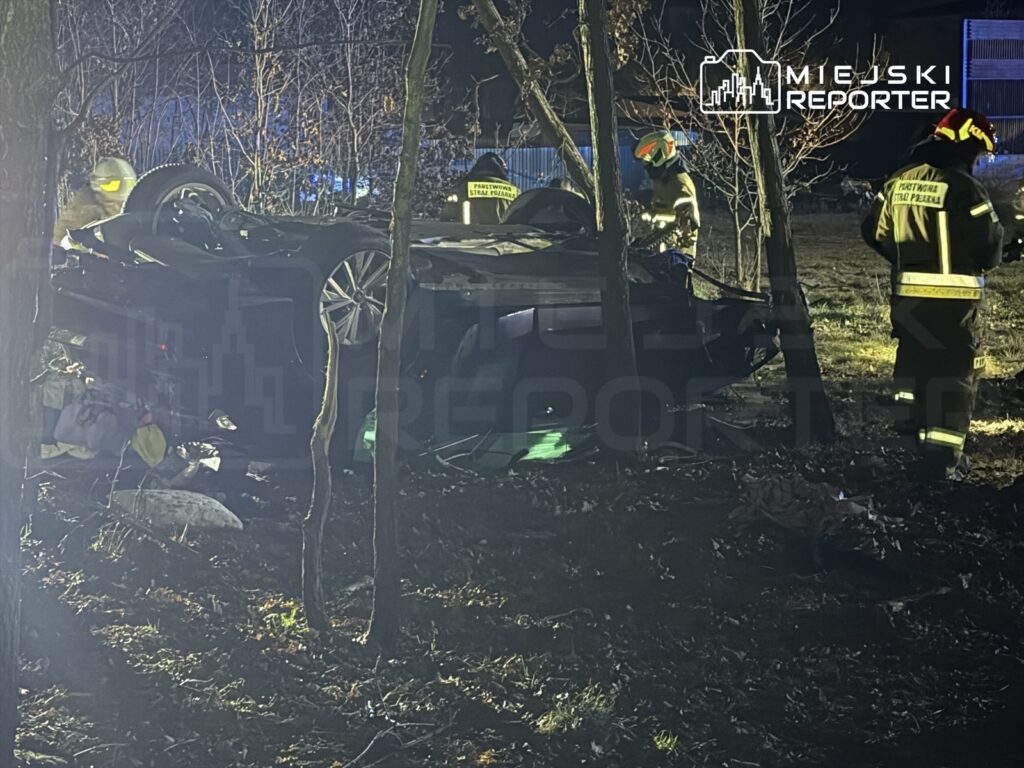 Ratownicy w mundurach przeszukują teren wokół przewróconego samochodu w lesie, w nocy.