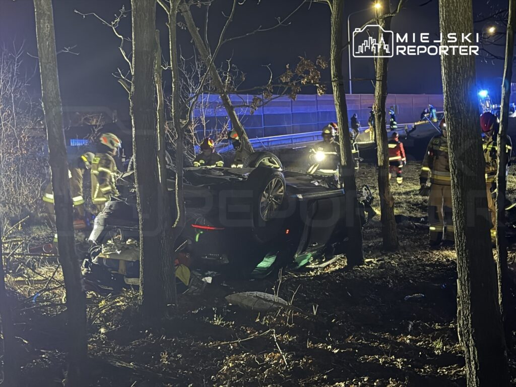 Grupa strażaków prowadzi akcję ratunkową przy przewróconym samochodzie w leśnym otoczeniu w nocy.