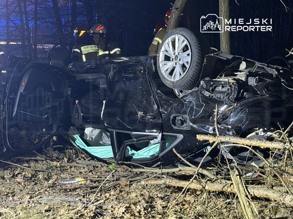 Samochód osobowy przewrócony na dach w lesie, otoczony przez strażaków w hełmach, w tle ciemne drzewa.