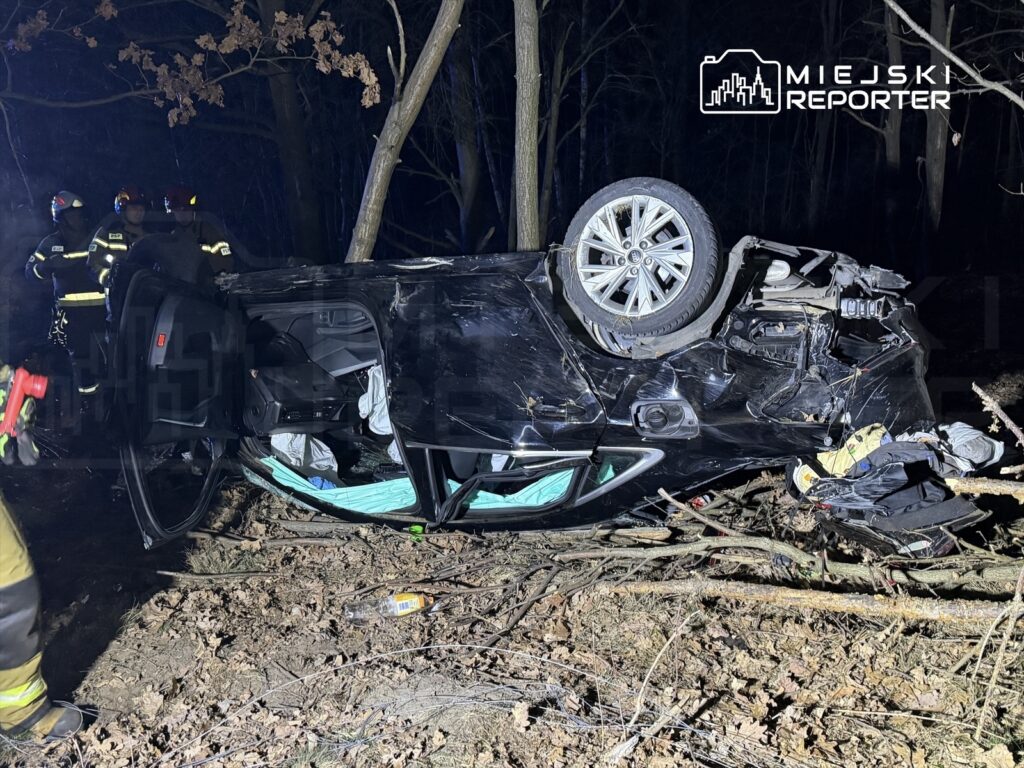 Strażacy pracują nad przewróconym samochodem osobowym w lesie, w nocy, w otoczeniu drzew i liści.