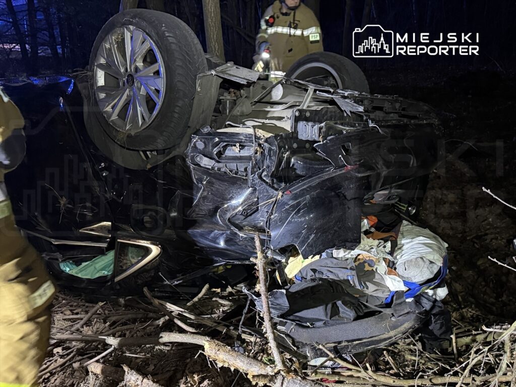 Samochód osobowy leży na dachu w lesie, otoczony gałęziami i zaroślami, obok pracują strażacy.