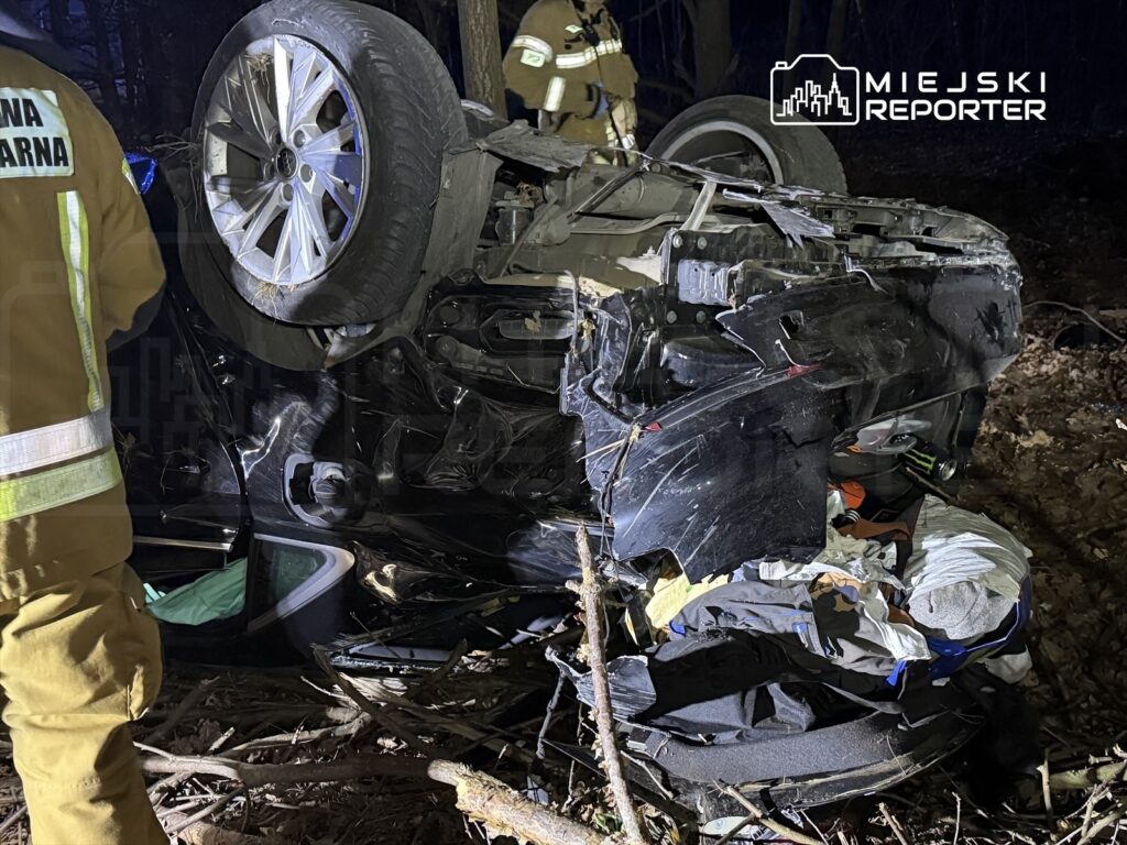 W lesie leży przewrócony samochód osobowy, a strażacy w mundurach przeszukują teren wokół pojazdu.