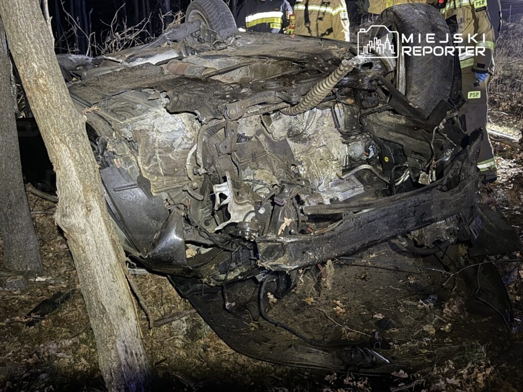 Zniszczony samochód leży na boku w lesie, otoczony przez strażaków oceniających sytuację.
