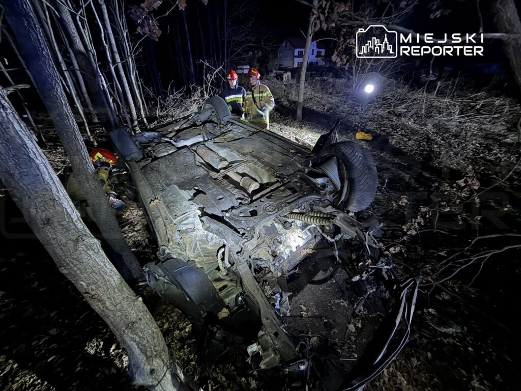 Strażacy w hełmach pracują przy przewróconym samochodzie w lesie, otoczonym drzewami i słabym oświetleniem.