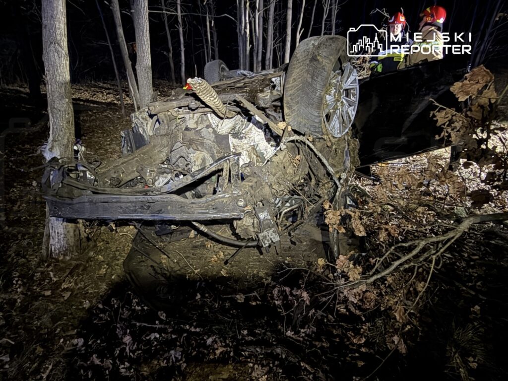 Samochód przewrócony na dach w lesie, otoczony drzewami i opadłymi liśćmi, z widocznymi uszkodzeniami.