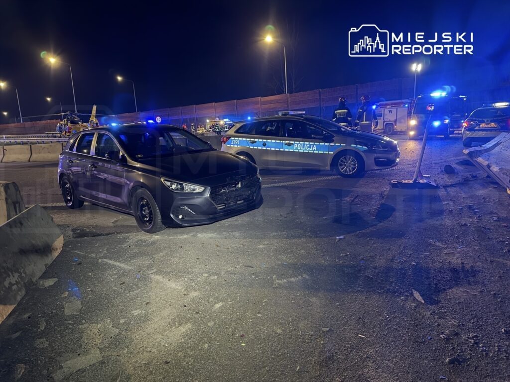 Czarny samochód osobowy stoi na poboczu drogi, obok policyjny radiowóz, w tle widać pojazdy ratunkowe i światła policyjne.