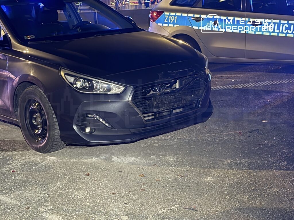 Czarny samochód osobowy z uszkodzoną przednią częścią stoi na parkingu, obok radiowozu policyjnego z niebieskimi światłami.