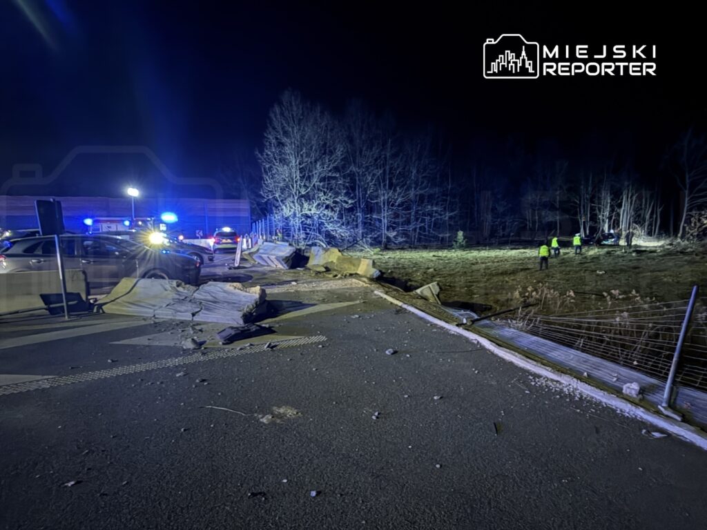 W nocy, przy drodze, zniszczona bariera ochronna, a obok pojazdy służb ratunkowych oraz osoby w kamizelkach odblaskowych.