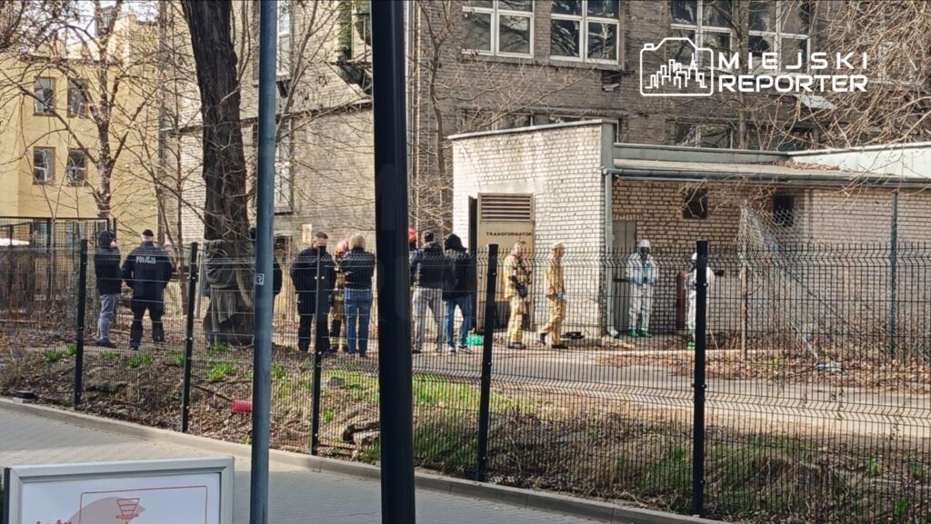 Grupa osób w mundurach policyjnych i strażackich stoi przed budynkiem z napisem „TRANSFORMATOR”, w tle drzewa.